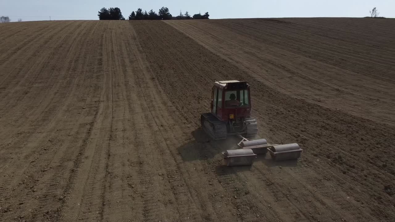Drone follow a red small tractor in rolling soil operation for seedbed preparation in uphill countryside agricultural farm
