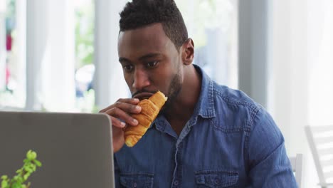 Hombre-De-Negocios-Afroamericano-Usando-Una-Computadora-Portátil-Y-Comiendo-Croissant-En-Un-Café