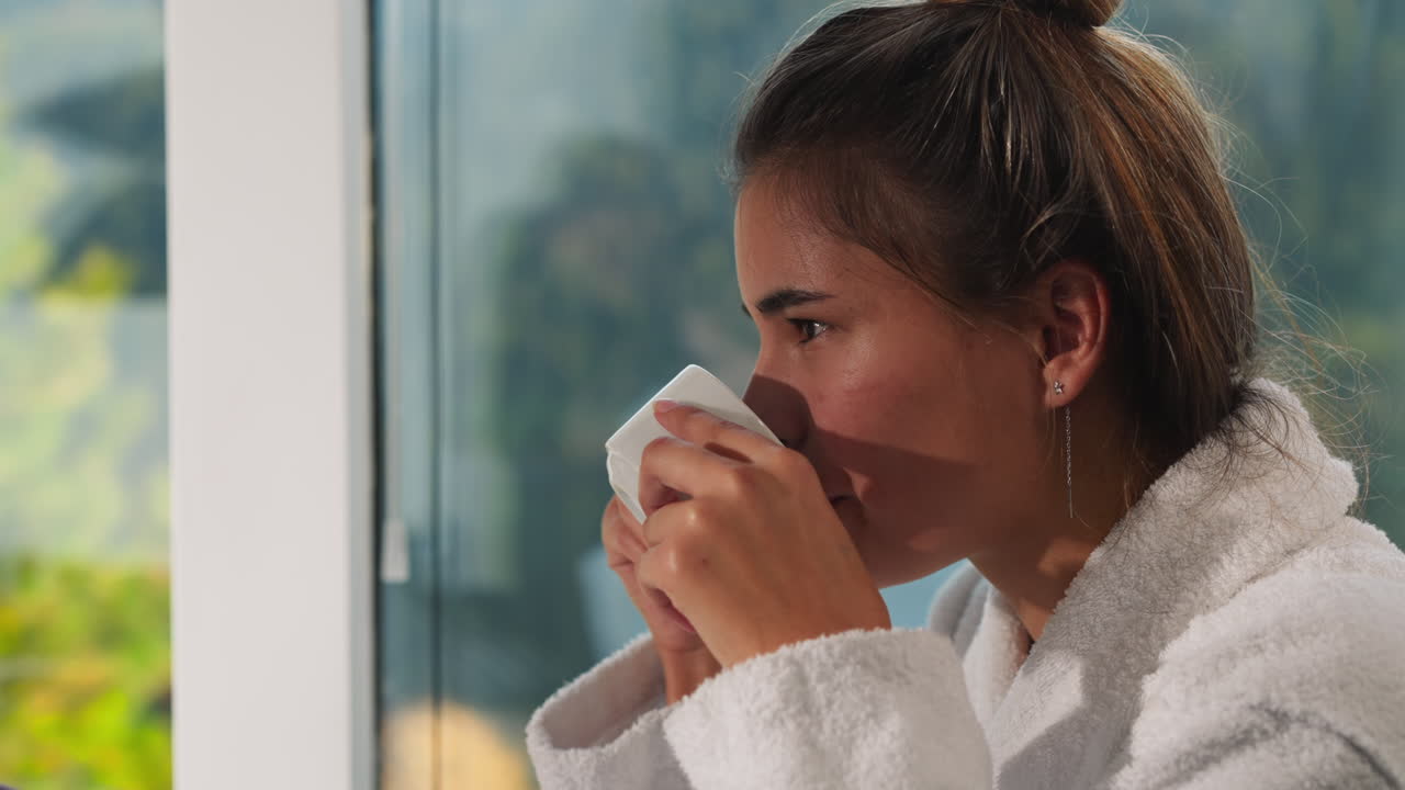 una mujer bebe café cerca de la ventana. una mujer muy positiva con túnica de terry toma un sorbo de té natural recién preparado en la cocina de casa. estilo de vida saludable