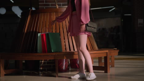 lady in a pink outfit places her shopping bags near a wooden sit out in a mall, she holds a black handbag and sits down, legs slightly crossed, with her left hand resting on the bench as she relaxes