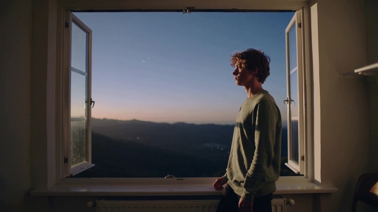 Entering man walking along window pausing at windowsill watching mountains in green top, copy space