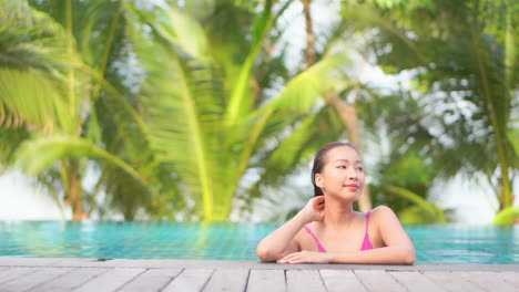 Una-Mujer-Joven-Y-Atractiva-En-Una-Piscina-Se-Inclina-A-Lo-Largo-Del-Borde-De-La-Piscina-Mientras-Mira-A-Su-Izquierda-Y-Derecha,-Con-Palmeras-En-El-Fondo