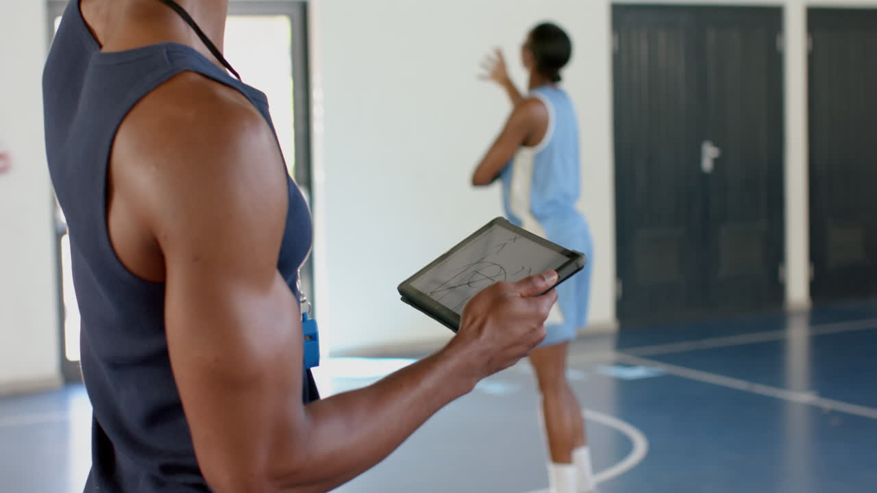 Coach holding tablet, observing basketball player practicing dribbling in gym
