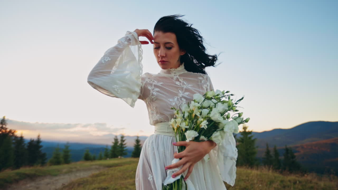 Woman in white dress and with bouquet in hands stands at the blowing wind. Lady touching her hair gently, closing her eyes in the rays of setting sun. Fantastic mountainous scenery at backdrop.