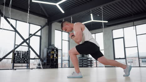 Guy doing warm up exercise includes side lunge and stretch in modern gym studio with large windows, wearing white tank top and black shorts, showcasing muscle activation, balance and focus