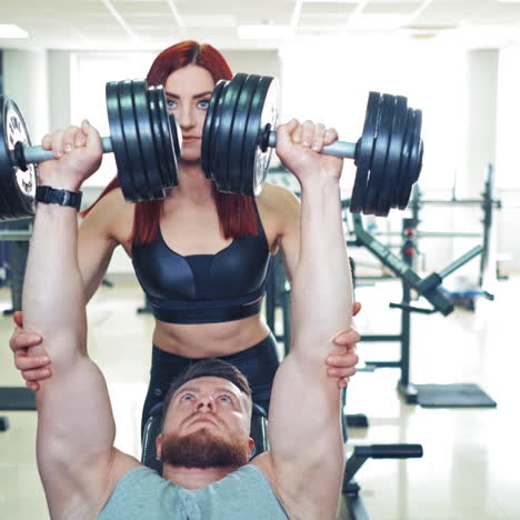strong man lifts dumbbells while working out with an instructor in sport club. attractive young woman is helping athletic man to do the exercise correctly. man finishes lifting heavy dumbbells.