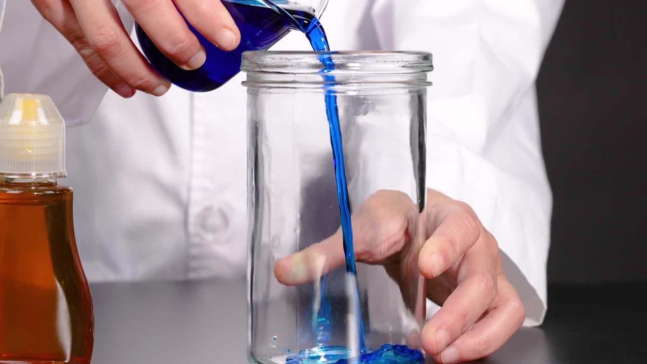 실험실 코트 를 입은 사람 이 검은색 표면 에 있는 갈색 병  에 있는 은 항아리 에 파란색 액체를 부어 놓는다