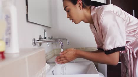 Mixed-race-gender-fluid-person-standing-in-bathroom-and-washing-face