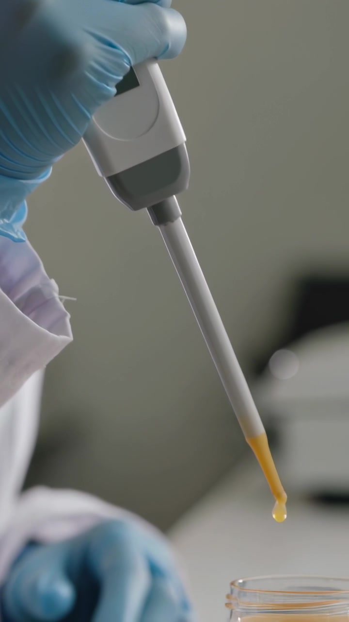 Video verticale: Un tecnico preme una pipetta, riempiendo un barattolo con liquido arancione su un banco, indossando camice e guanti.
