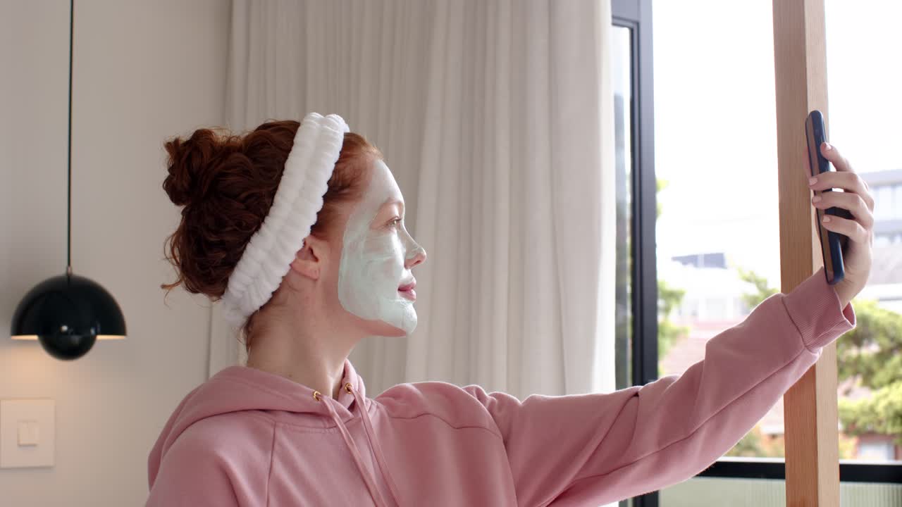 Mulher adulta segurando um smartphone, gravando a rotina de skincare e posando com máscara facial perto da janela