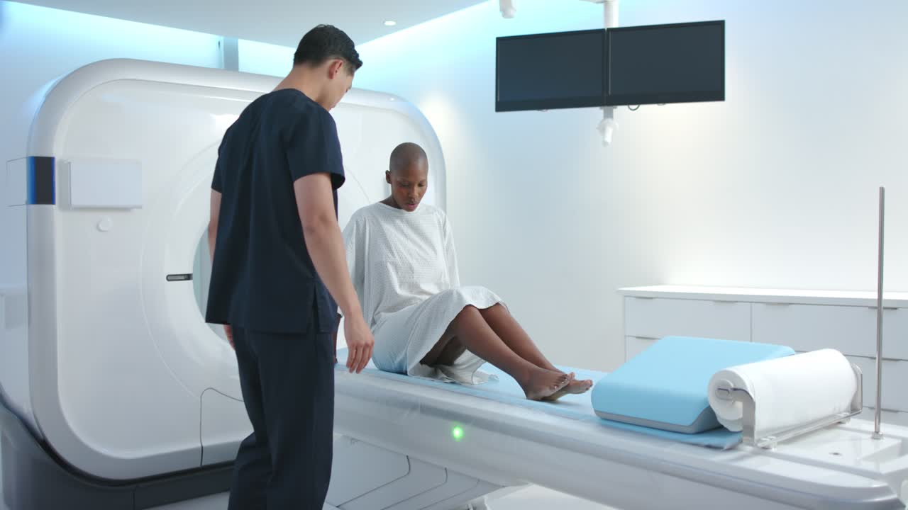 Tecnico in divisa medica che guida delicatamente una donna in camice ospedaliero che si sistema sul lettino della risonanza magnetica per la preparazione della scansione.