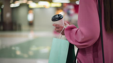 back view of woman in pink dress holding coffee cup and shopping bags while walking through a brightly lit mall, stores and chairs with tables can be seen with blurred background and mall lights