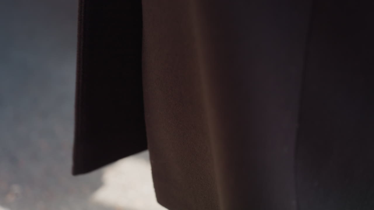 섬세한 디테일이 돋보이는 울 롱 코트가 밑단에서 흔들리고, 햇빛을 받아 반짝이는 스냅 버튼, 부드러운 벤치가 배경에 있으며, 길거리 분위기, 미니멀리즘 패션 바이브, 차분한 색상과 부드러운 바람이 원단을 움직이는 모습이 보여요.