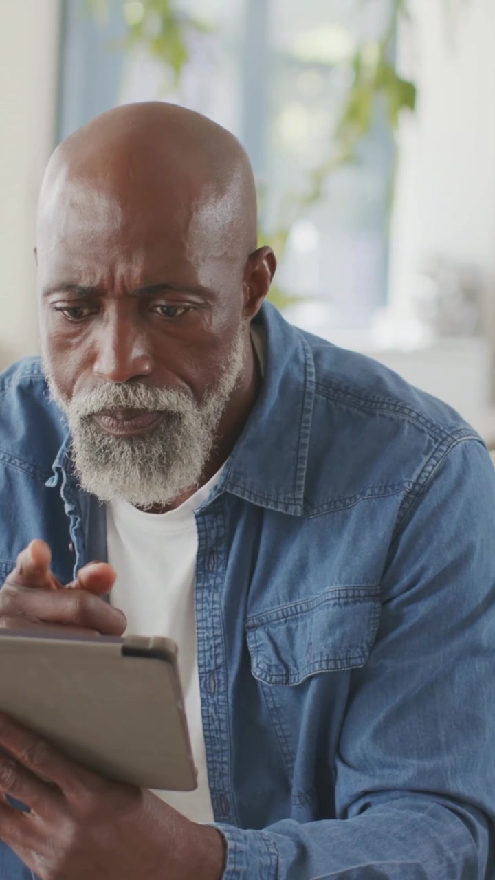 Vertical video: Holding tablet, man wearing denim shirt entering card details to pay in living room