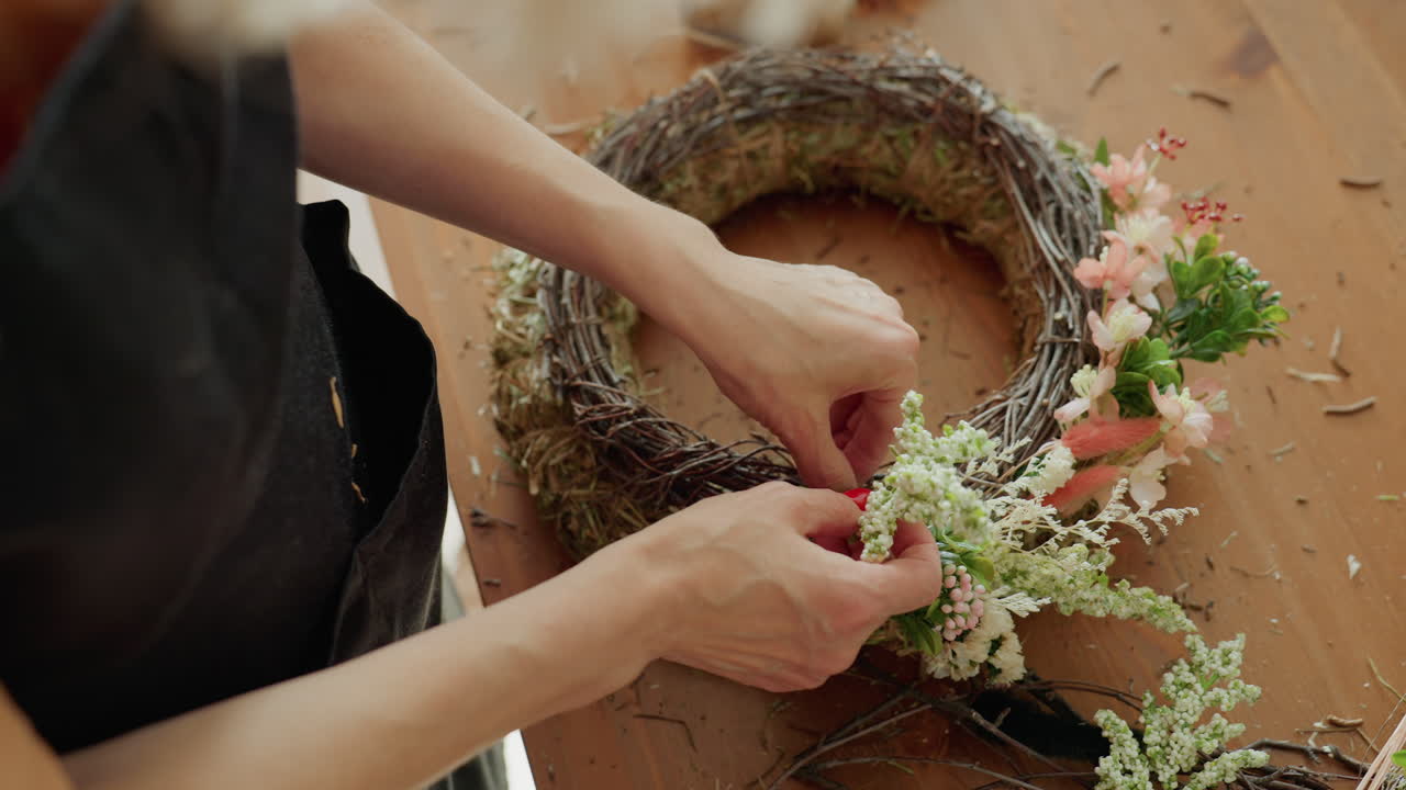 Close up of florist hand arranging white flowers and green foliage on rustic twig wreath over wooden table during creative floral decoration process with natural elements and seasonal blossoms