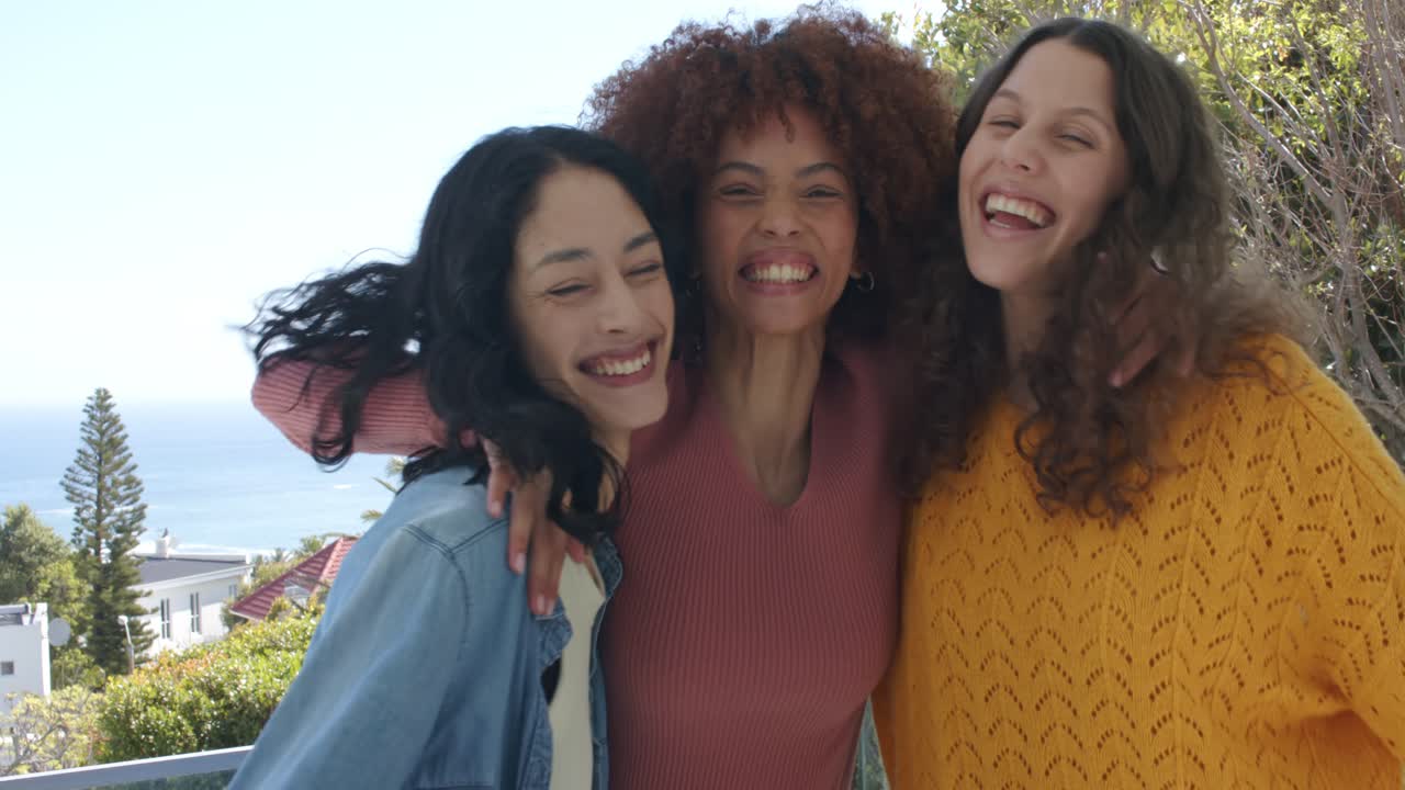 Amigas diversas dançando em varanda de vidro, entrando, se abraçando e rindo com vista para o oceano