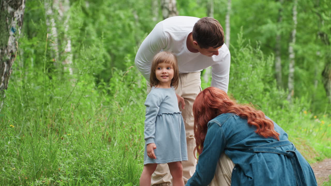 Una madre premurosa si inginocchia per allacciare il sandalo della sua giovane figlia su un sentiero nel bosco, mentre il fratello è in piedi accanto a loro, circondati da alberi verdeggianti; un caloroso momento familiare che mostra affetto e attenzione genitoriale all'aria aperta.