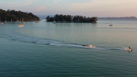 toma aérea panorámica que captura la actividad de los deportes acuáticos, paseo en bote banana tirando detrás de una moto de agua con un hermoso punto de referencia de la casa de luz del muelle de perdana en el fondo al atardecer, isla langkawi