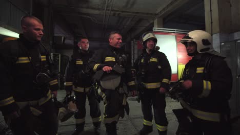 group of five firefighters wearing full equipment in smokey area. relaxed chatting after work well done. fire trucks in the background. name tag translation is fireman and model's last name.