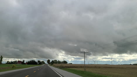 POV-shot-of-a-motor-vehicle-travelling-along-a-remote-country-road-passing-a-series-of-wind-turbines,-above-ominous-dark-grey-rain-clouds-build,-Toronto,-Canada