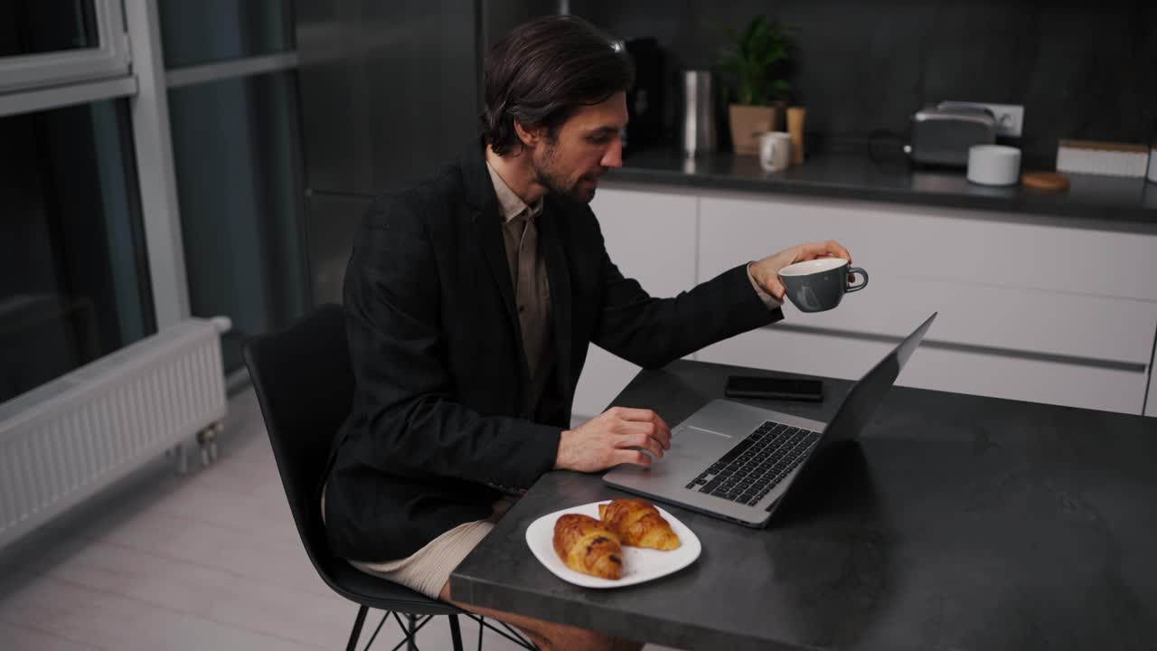 A confident brunette man with stubble in a black jacket works remotely and communicates using a laptop while sitting on a chair in beige shorts in the evening in a modern apartment