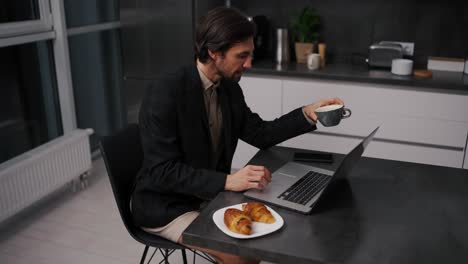 A-confident-brunette-man-with-stubble-in-a-black-jacket-works-remotely-and-communicates-using-a-laptop-while-sitting-on-a-chair-in-beige-shorts-in-the-evening-in-a-modern-apartment