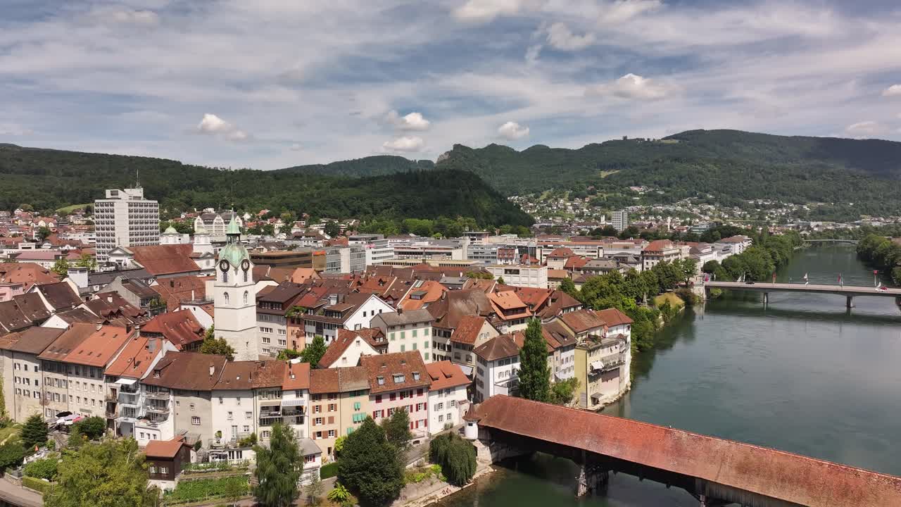 Olten town drone view with red-roof old town buildings, covered wooden bridge and Aare river flowing through Canton Solothurn, Switzerland, lush green hills and sunny skies