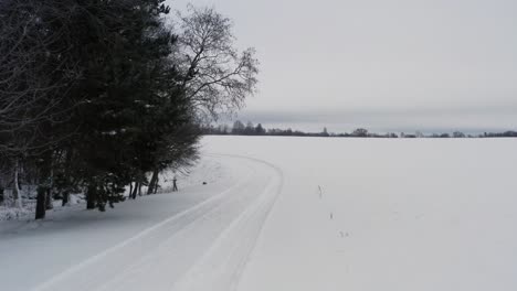 Kleine-Verschneite-Landstraße-Mit-Autoreifenspuren-In-Der-Nähe-Von-Baum-Und-Fluss