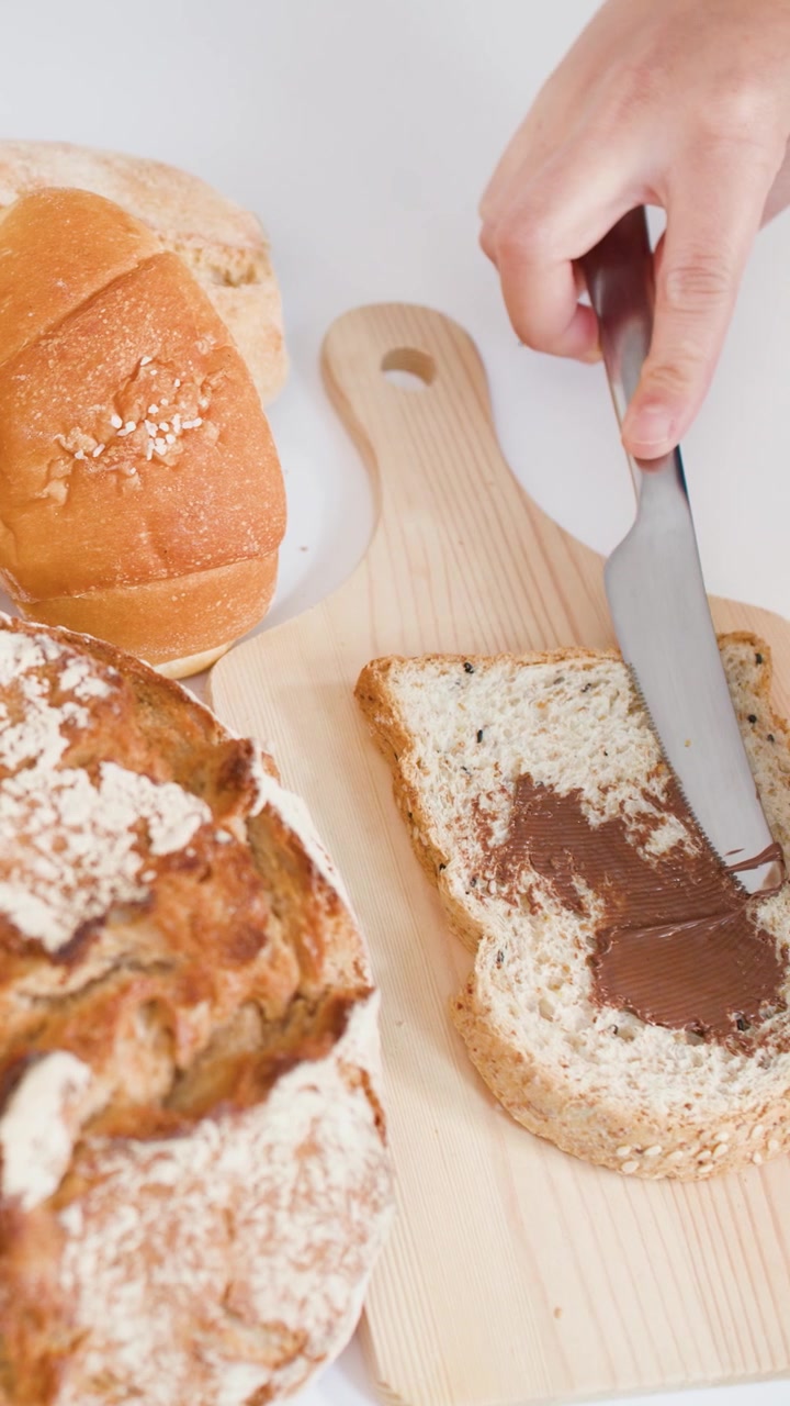 Hand spreads chocolate cream on bread slice beside croissants and sourdough on wooden board.