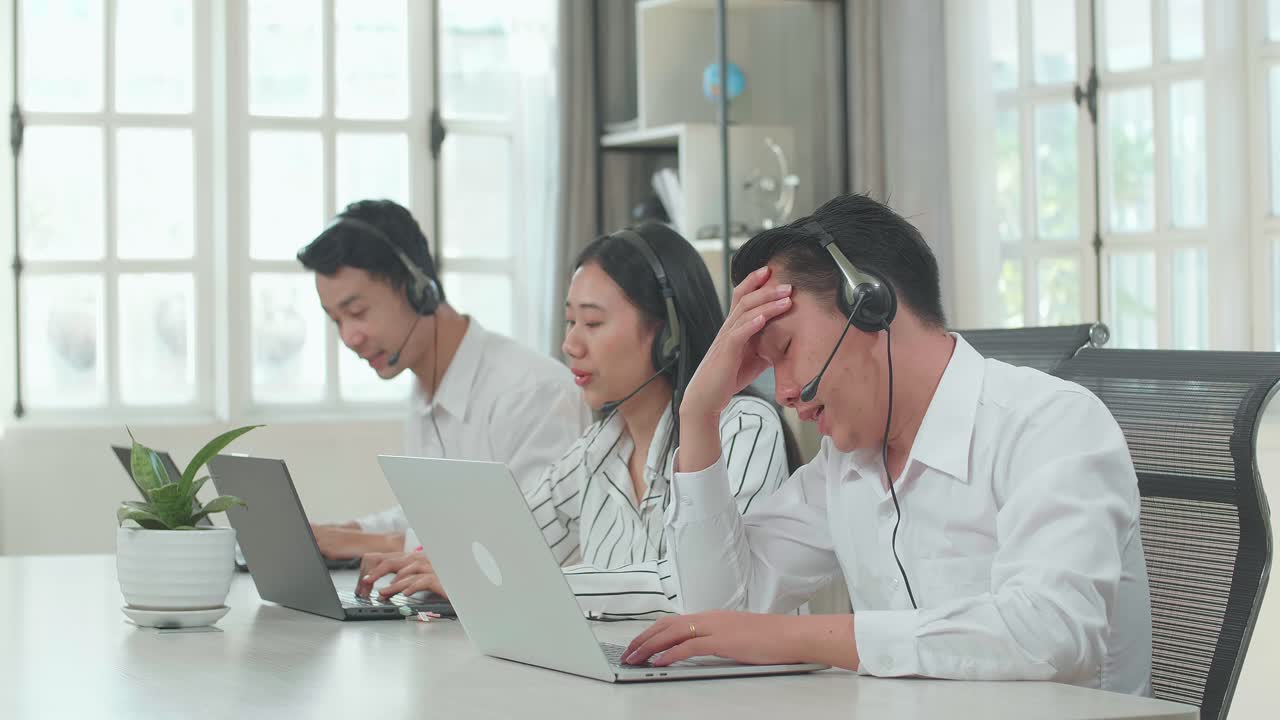 A Man Of Three Asian Call Center Agents Wearing Headset Is Tired Due To Working While Two Of His Colleagues Are Speaking And Typing During The Call With Customer At The Office
