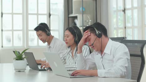 a man of three asian call center agents wearing headset is tired due to working while two of his colleagues are speaking and typing during the call with customer at the office