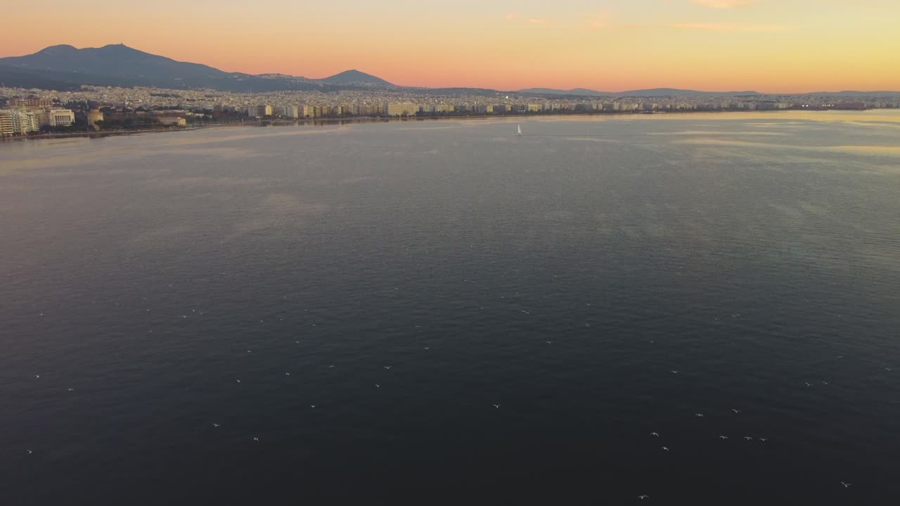 Sunset Flight Over Thessaloniki Waterfront: Aerial Drone View Captures Birds Gliding Above Tranquil Aegean Waters, Near Vibrant Aristotle Square – Scenic Greece Coastal Cityscape in Golden Light