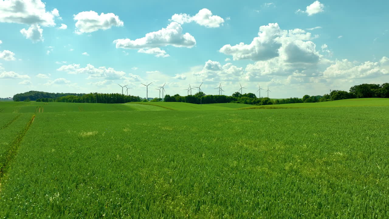 Vue aérienne de vastes champs verts avec des éoliennes et un ciel bleu éclatant parsemé de nuages cotonneux.