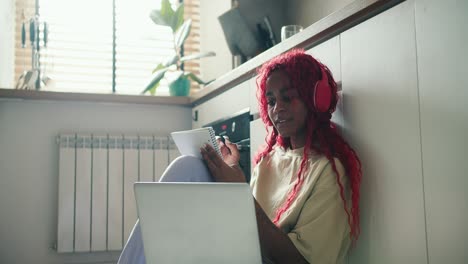 Una-Chica-Afroamericana-Con-El-Pelo-Rizado-Rojo-Estudiando-En-El-Suelo-De-La-Cocina-Con-Un-Portátil,-Tomando-Notas