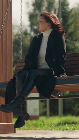 joven con abrigo negro y botas de cuero en un columpio de madera en el parque, el cabello balanceándose con el viento, rodeada de flores y vegetación durante un día soleado