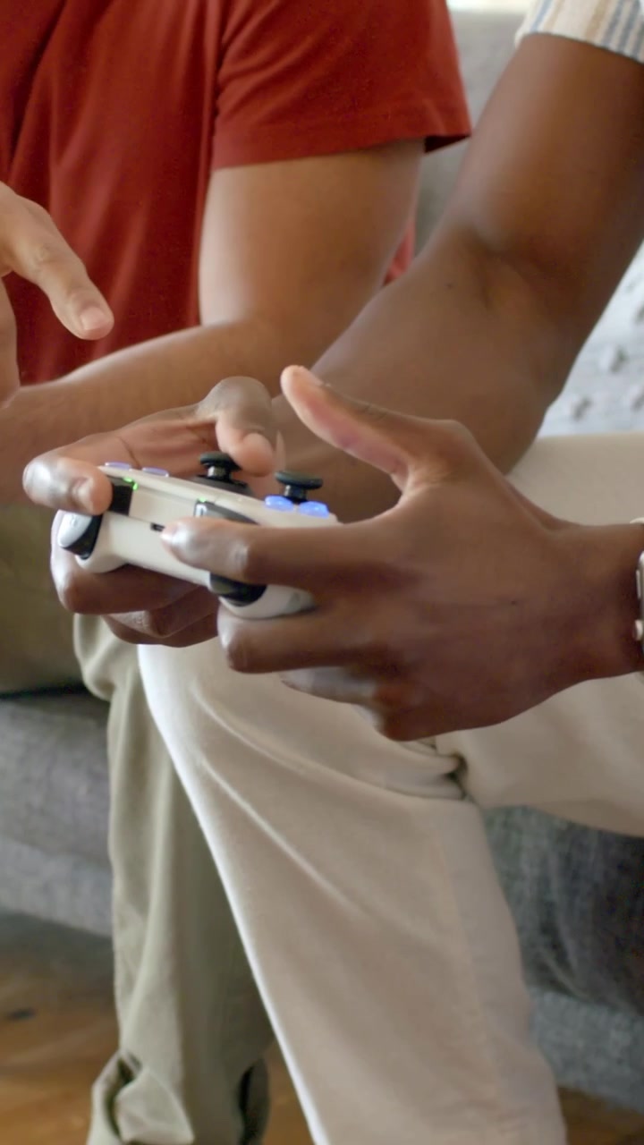 Video verticale: Due uomini con magliette a righe color ruggine che iniziano una partita, premendo i gamepad sul divano con degli snack.