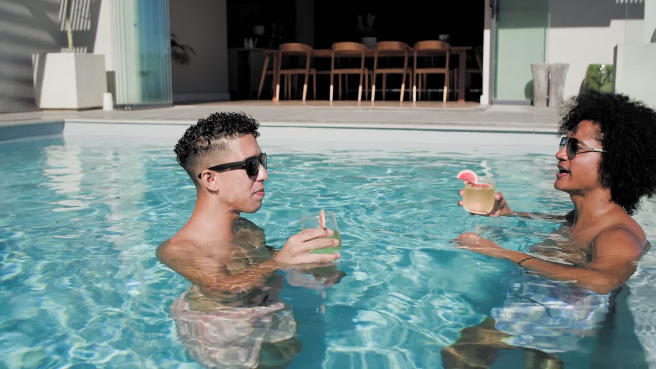 Afroamerikanische Männer stoßen mit Cocktails an, unterhalten sich und zeigen vom Dachpool aus auf das Meer.