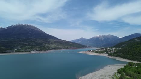 Crisp 4K drone footage of snowy mountains surrounding Lac de Serre Ponçon, focusing on Savines Le Lac Bridge and Mont Guillaume. Captured in the French Alps, May 2025, showcasing natural beauty