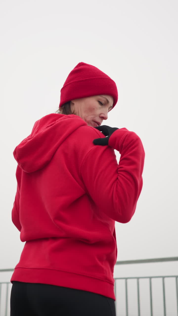vista lateral de una mujer realizando un estiramiento de la mano girando el brazo al aire libre en una rutina de fitness, vestida con ropa atlética, suelo cubierto de nieve, árboles helados, banco, barandilla de hierro en un parque sereno