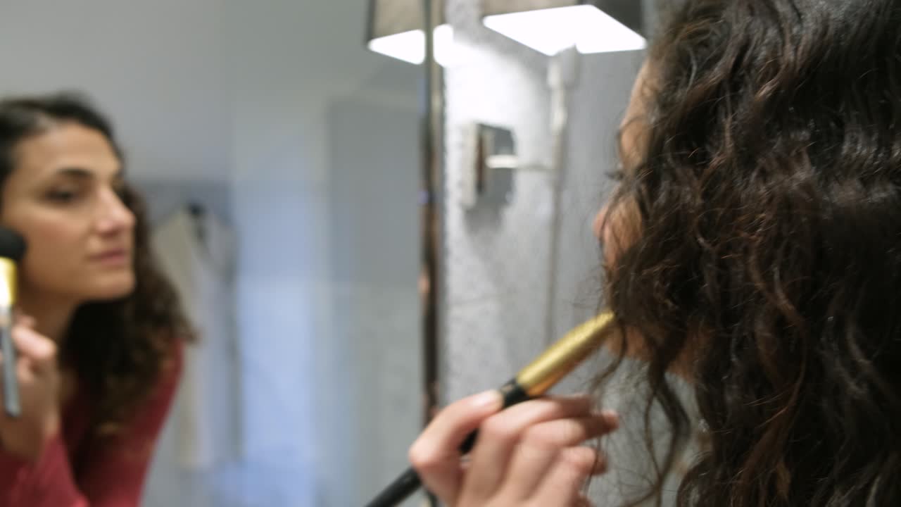 Una hermosa mujer morena de Oriente Medio aplicándose polvos de maquillaje con una brocha grande en los pómulos mientras se mira en el espejo.