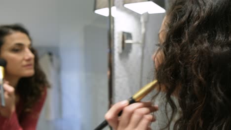 una hermosa morena del medio oriente aplicando polvo de maquillaje con un cepillo grande en los pómulos mientras se mira en el espejo