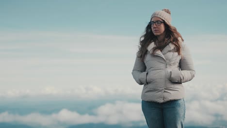 slow motion of a young woman with long brown hair and glasses standing on top of a beautiful mountain landscape on a windy and cloudy day