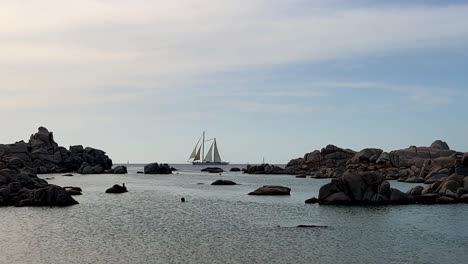 Double-mast-sailing-ship-crosses-the-scene-at-Cala-della-Chiesa-on-Lavezzi-island-in-Corsica-with-people-bathing,-France