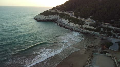 Costa-garraf-coastline-with-waves-hitting-the-shore-near-an-old-cement-factory-at-sunset,-aerial-view