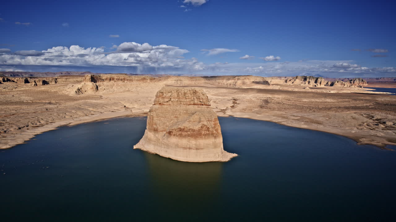 Cinematic drone shot soaring over Lake Powell, focusing on a massive solitary rock framed by the surrounding desert terrain.