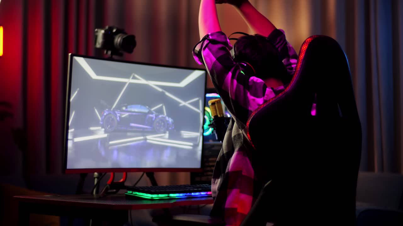 Back View Of Asian Boy Streamer Stretching While Playing Car Racing Game Over Network On Personal Computer. Live Stream Video Game, Desk Illuminated By Rgb Led Strip Light