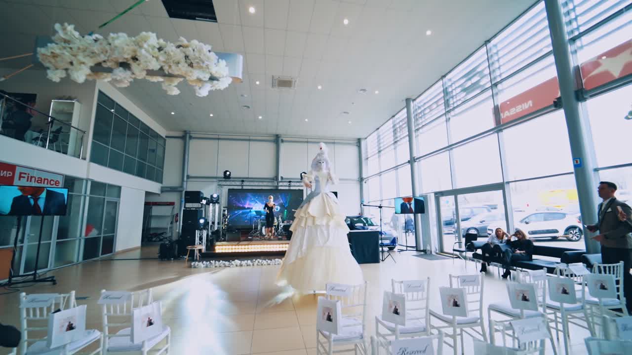 Preparation for the show. Tall woman in mask and white dress moving in the hall. Singer is singing on stage. Empty chairs in the room.