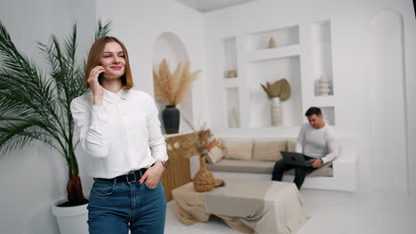 slim positive lady in white shirt speaks on the phone. man sitting at backdrop holding a laptop.
