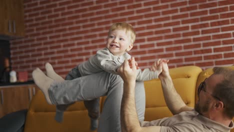 Un-Hombre-Alegre-Sosteniendo-A-Un-Niño-Pequeño-Saltando-Divirtiéndose-Juntos-En-El-Sofá-De-La-Sala-De-Estar