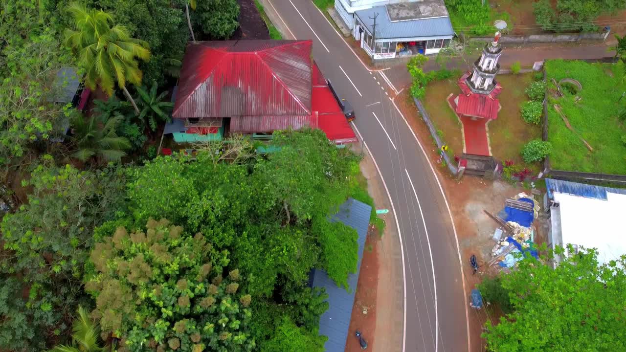 Aerial top-down shot tracking a winding rural road and pedestrian crossing through a dense tropical village with colorful red and blue roofs and a small roadside shrine tower